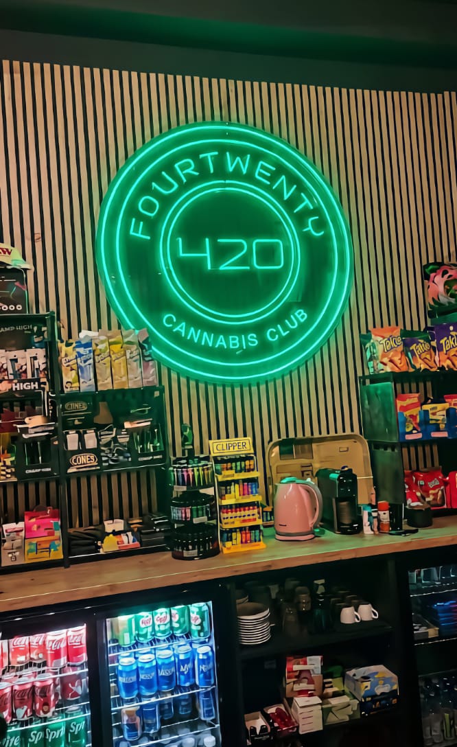 Un'insegna al neon verde brillante con il logo del cannabis club Four Twenty di Valencia, esposta sopra un bancone del bar ricco di snack, bevande e accendini Clipper.