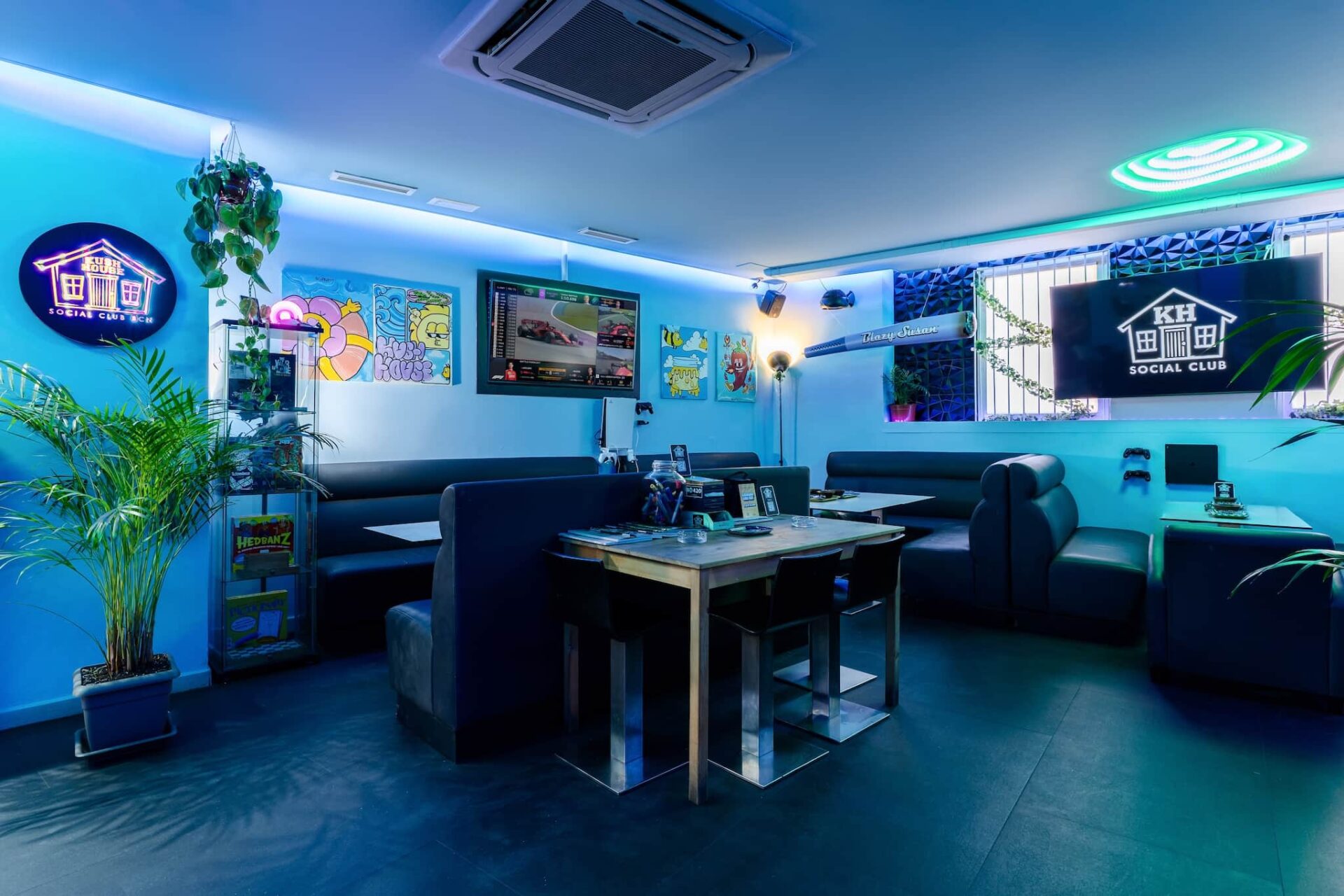 Interno del Kush House Social Club a Barcellona, con illuminazione blu soffusa, sedute a divanetto, schermi per il gaming, murales colorati e piante in vaso