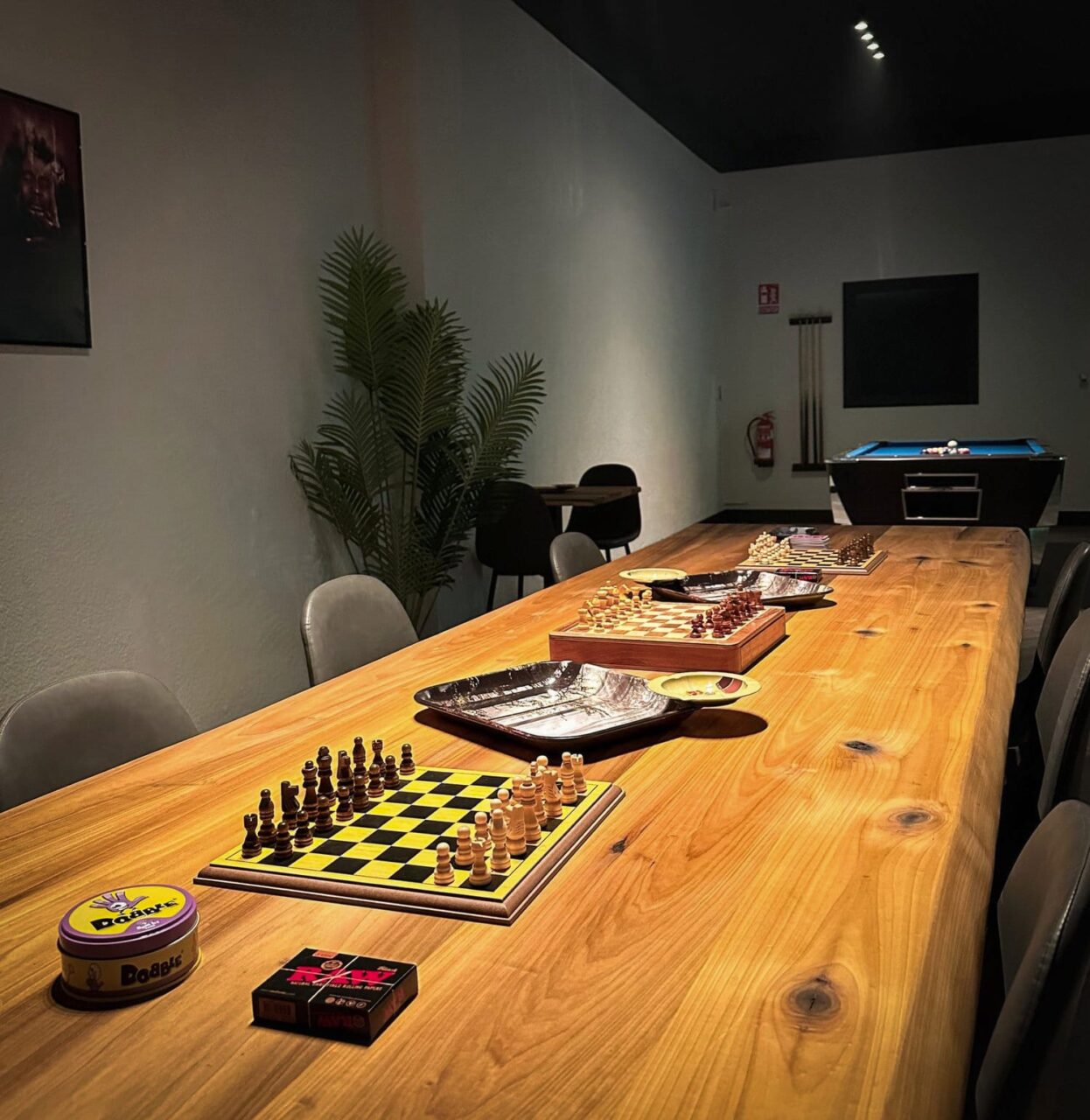Un lungo tavolo in legno allestito con scacchiere, giochi da tavolo e accessori per fumatori presso il cannabis club Four Twenty di Valencia, con un tavolo da biliardo visibile sullo sfondo.