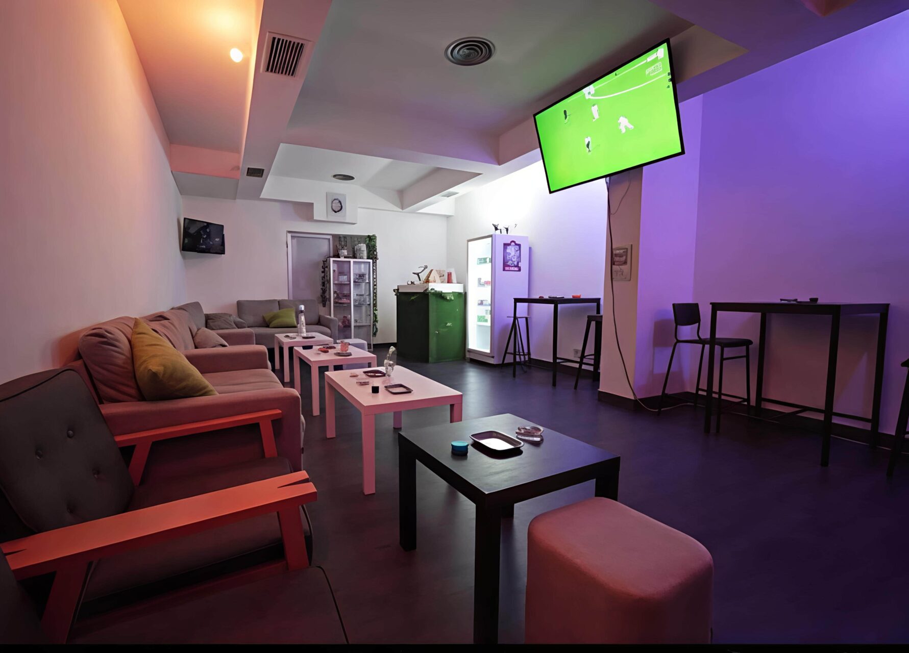 Area lounge nel Valhalla cannabis club a Madrid con posti a sedere, tavoli alti, un frigorifero e una TV che trasmette una partita di calcio.