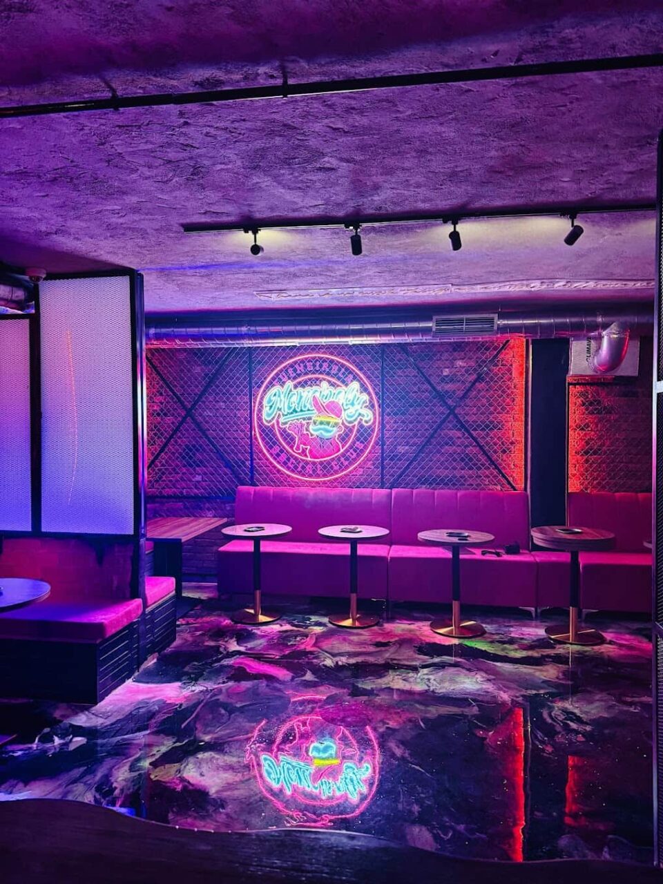 Area lounge del Monopoly Social Club a Malaga, con illuminazione ambientale viola, muro di mattoni, divani rosa imbottiti e pavimento riflettente.  